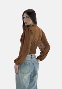Blusa marrone, trasparente, a maniche lunghe con vita arricciata, abbinata a jeans chiari, a taglio morbido. Tessuto liscio con design a spalla gonfia.