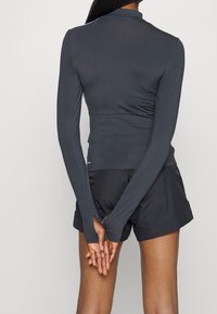 Donna in piedi con le mani incrociate dietro la schiena, che indossa un top aderente a maniche lunghe di colore grigio scuro e pantaloni corti sportivi neri.