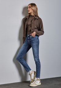 Giacca marrone con bottoni sopra una maglietta marrone aderente, abbinata a jeans skinny blu con borchie laterali e sneakers beige e bianche.