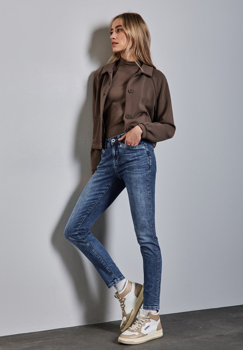 Giacca marrone con bottoni sopra una maglietta marrone aderente, abbinata a jeans skinny blu con borchie laterali e sneakers beige e bianche.