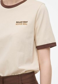 T-shirt in cotone beige con colletto e polsini a coste marroni. Presenta la scritta "ROUGE EDIT" in marrone e oro sul davanti.