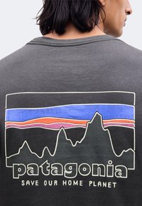 Άτομο που φοράει σκουρόγκρι μπλουζάκι Patagonia με πολύχρωμο λογότυπο βουνού και κείμενο "Save Our Home Planet" στην πλάτη.