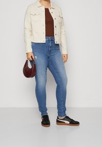Hellbeige Jeansjacke, braunes Rippoberteil, abgenutzte blaue Skinny-Jeans und schwarze Sneakers mit Gummisohlen. Hält eine burgunderfarbene Handtasche.
