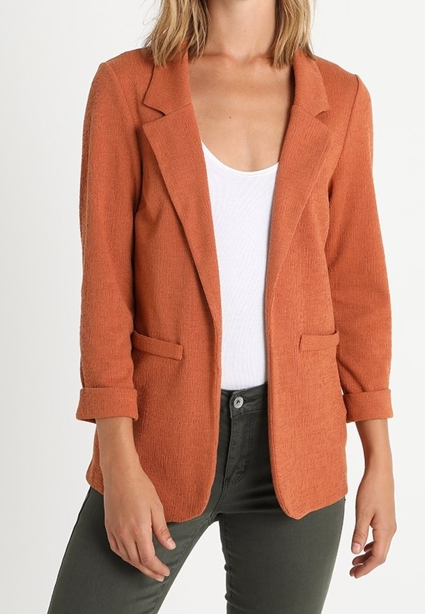 Femme portant un blazer texturé ouvert couleur orange brûlé sur un haut blanc à encolure dégagée et un pantalon vert foncé.