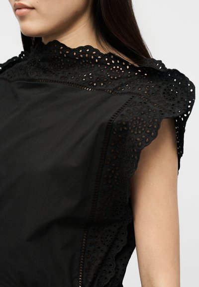 Γυναίκα με μαύρο αμάνικο τοπ με περίτεχνη δαντέλα με λουλούδια στο ψηλό λαιμό και στους ώμους, σε απλό φόντο.