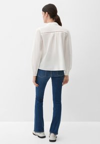 Donna con capelli scuri raccolti in una coda di cavallo, indossa una camicia bianca a maniche lunghe, jeans blu e sneakers bianche, in piedi contro uno sfondo bianco.