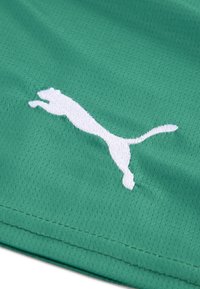 Hvid Puma-logo broderet på grønt åndbart stof med synlig syning langs kanten af materialet.