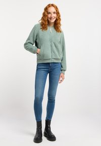 Mintgroene, fuzzy, ritsjacket met zijzakken, geribbelde manchetten en zoom, gecombineerd met blauwe skinny jeans en zwarte enkellaarsjes.