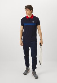 Tengerészsapkás póló piros gallérral, kék vízszintes csíkokkal. Sötét tengerészkék melegítőnadrággal és szürke sportcipővel viselve, teniszezőütőt tartva.