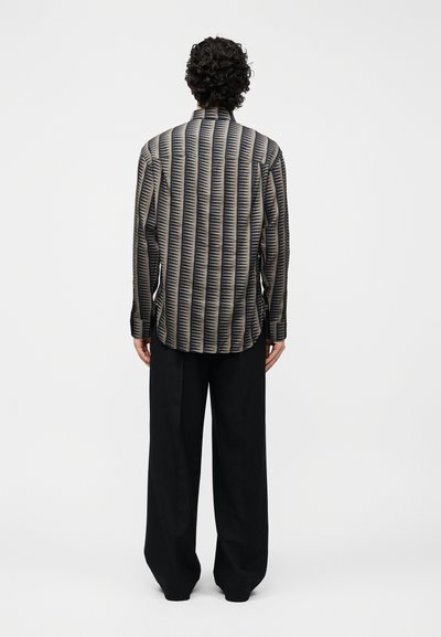 Άτομο με σγουρά μαλλιά που φοράει μπλούζα με σχέδια μακρυμάνικη και φαρδύ μαύρο παντελόνι, στέκεται με την πλάτη προς το μέρος μας μπροστά από απλό φόντο.