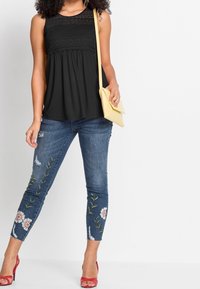 Svart ärmlös topp med spetsdetaljer, i kombination med slitna blå jeans med blommönster och fransad fåll. Gul handväska och röda klackar.