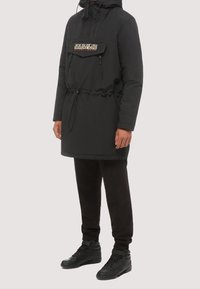 Chaqueta negra de manga larga con cordón en la cintura, capucha y un bolsillo delantero con logo. Combinada con pantalones negros y zapatillas.