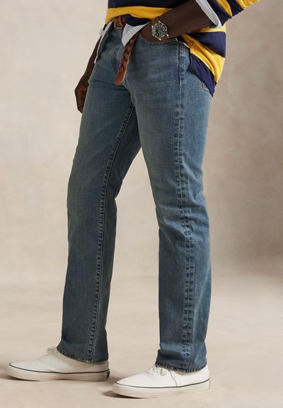 Person iført blå jeans, hvide sneakers, et flettet brunt bælte og en langærmet, stribet skjorte i navy og gul.