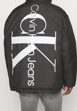 Doudoune noire avec grand logo blanc Calvin Klein Jeans dans le dos, portée par une personne en jean gris, de dos.