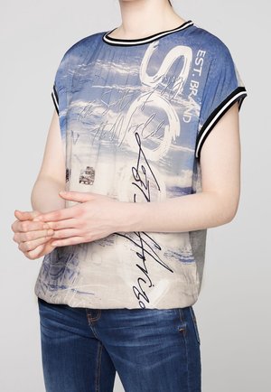 Osoba w niebiesko-beżowej koszuli z krótkim rękawem z nadrukiem, z biało-czarnymi paskowanymi kołnierzykami, w niebieskich dżinsach, trzymająca ręce złożone razem.