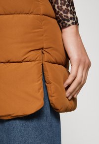 Brun puffer vest med quiltet design, sideslitt og en hånd som hviler på stoffet. Bær på denim, som viser tekstur og fargekontrast.