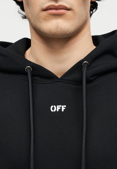 Tuvplāns cilvēkam, kurš valkā melnu džemperi ar kapuci, uz kura krūtīm baltā krāsā redzams uzraksts "OFF", redzamas auklas un daļēji seja.