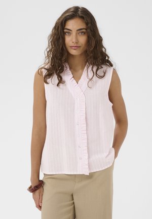 Vrouw met krullend bruin haar, gekleed in een mouwloos lichtroze gestreept blouse met ruchtrand en beige broek, staand tegen een eenvoudige achtergrond.