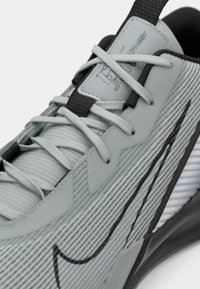 Tuvu pelēks sporta apavus ar gaiši pelēkām šņorēm, melnu iekšējo odere, teksturētu auduma augšdaļu un melnu Nike swoosh logotipu sānos.