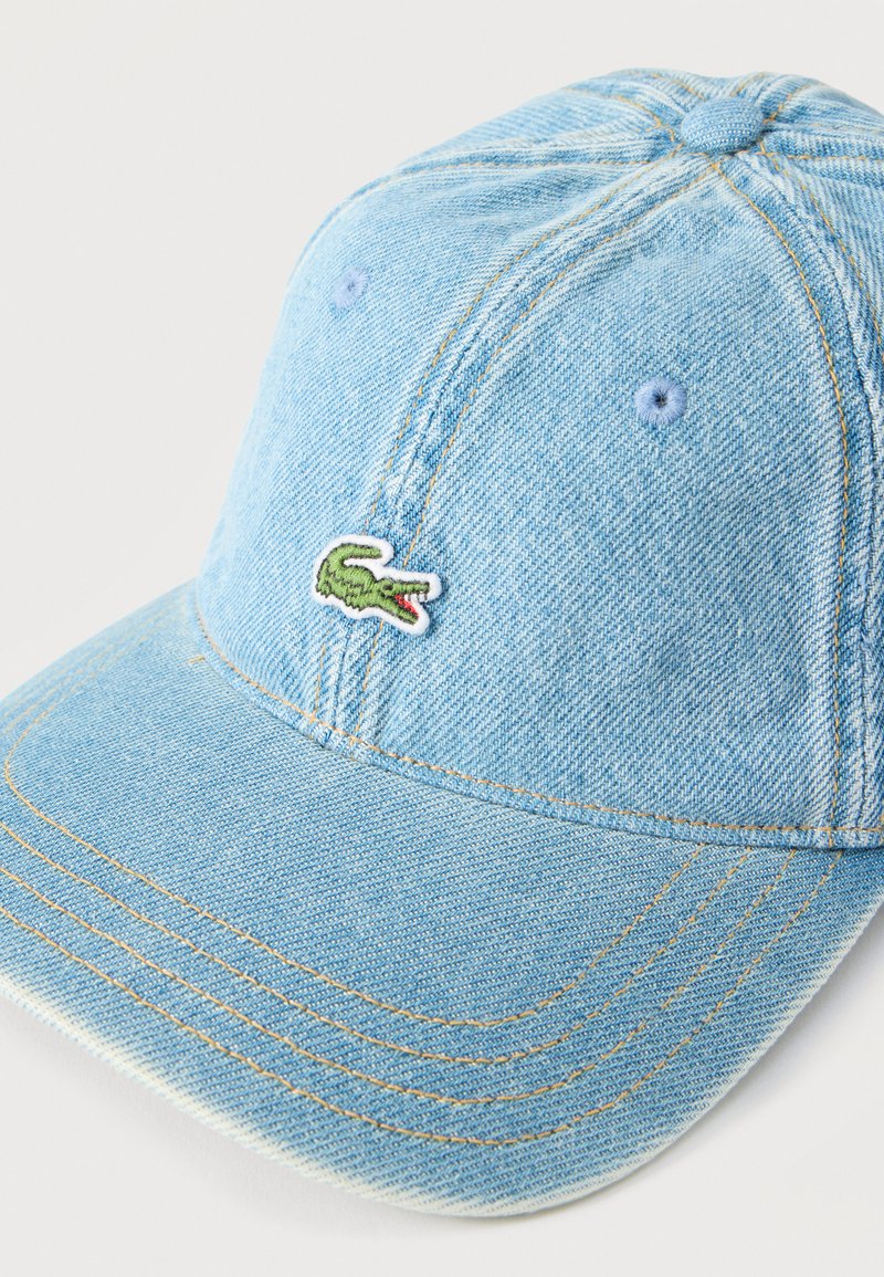 Cappellino da baseball in denim azzurro chiaro con ricamo a logo di coccodrillo verde sulla pannello frontale e cuciture gialle visibili.