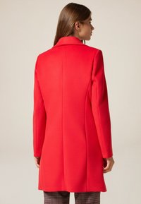 Abrigo de lana rojo con un corte entallado. Presenta un corte recto, cuello con muescas y tejido texturizado. Acabado liso, sin cierres visibles.