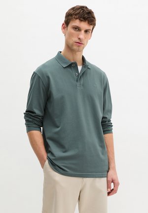 Young man with short hair wearing a muted green long-sleeve polo shirt and beige pants, standing with one hand in pocket.