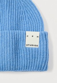 Világoskék kötött sapka bordázott textúrával, fehér címkével a peremén, melyen fekete betűkkel "CPH STUDIOS" felirat olvasható.