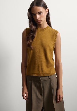 Femme aux longs cheveux foncés portant un pull sans manches jaune moutarde et un pantalon ample marron foncé, debout devant un fond clair uni.