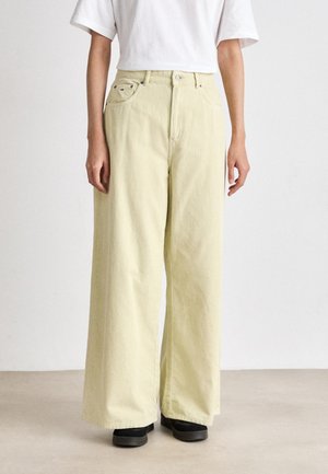 Pantalons en velours côtelé jaune clair à jambe large avec poches avant et taille haute, associés à une chemise blanche à manches courtes et des chaussures noires.