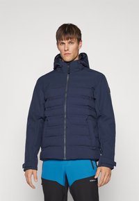 Μπλέ ναυτικό αδιάβροχο μπουφάν με οριζόντιο ραβδωτό σχέδιο, μπροστινό φερμουάρ, αποσπώμενη κουκούλα και πλεκτούς μανικέτες, φορεμένο πάνω από φωτεινά μπλε παντελόνια.