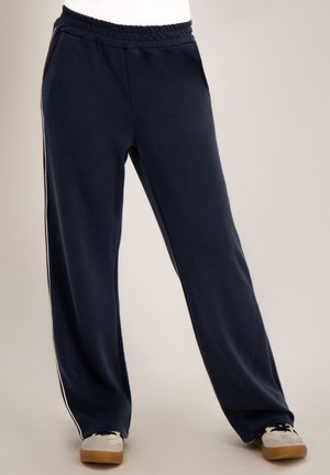 Marineblauwe broek met wijde pijpen, elastische tailleband en strepen aan de zijkant, gedragen met witte en beige sneakers op een neutrale achtergrond.