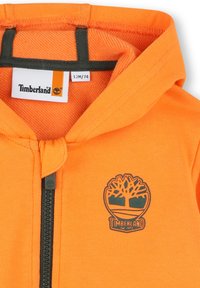 Sweat à capuche orange avec fermeture éclair, arborant un logo Timberland vert et un tissu texturé. L'étiquette de taille indique 12M/74.