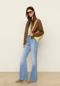 Veste en daim marron, haut blanc, jeans flare bleu clair avec fentes sur les côtés, et chaussures beiges. L'accessoire comprend un sac à épaule jaune.