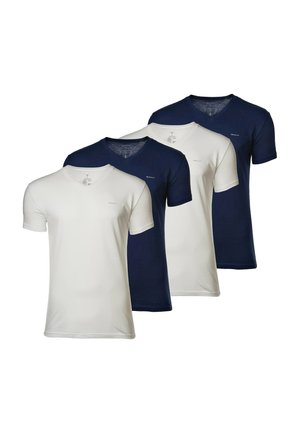 Młody mężczyzna w granatowym T-shircie, białych bokserkach i białych skarpetkach, lekko podnosi koszulkę, aby odsłonić gumkę.