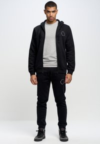 Schwarze Zip-up-Trainingsjacke über einem grauen T-Shirt, kombiniert mit schwarzen Jogginghosen mit einem runden Logo. Das Material wirkt weich und strukturiert.