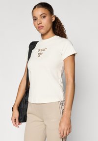 Camiseta de manga corta color crema, hecha de tejido suave, con un logo marrón impreso en el centro. La modelo sostiene un bolso negro y lleva pantalones beige.