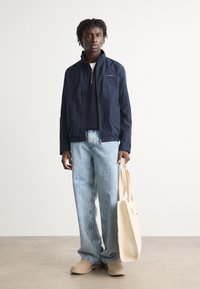 Veste marine, jeans bleus ajustés, chaussures beiges et un sac fourre-tout de couleur claire. La veste a une fermeture éclair à l'avant et un col haut.