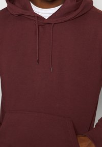 Bordowy hoodie z kieszenią kangurką, teksturowany materiał, kaptur ze sznurkiem i ściągacze przy mankietach. Posiada dwa nałożone białe wykończenie przy okrągłym dekolcie wewnętrznym.