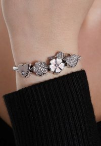 Silver bracelet featuring heart, flower, and gem charm designs. Textured finish with enamel accents. Worn on a wrist with black fabric.