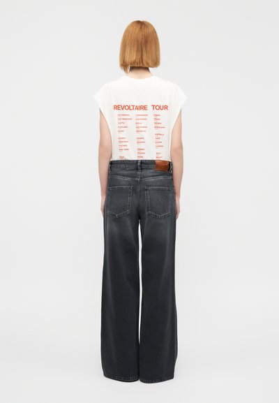 Λευκή μπλούζα με στάμπα "REVOLTAIRE TOUR" σε κόκκινο στο πίσω μέρος, σε συνδυασμό με μαύρο ψηλόμεσο, φαρδύ τζιν παντελόνι που διαθέτει καφέ δερμάτινο ταμπελάκι.
