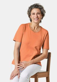 Korte mouwen oranje t-shirt, ronde halslijn, gemaakt van zachte katoen. Model zit op een houten stoel, draagt een witte broek, neutrale achtergrond.
