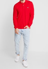 Polo shirt rosso a maniche lunghe con collo e un piccolo logo, abbinato a jeans di denim blu chiaro e sneakers bianche.