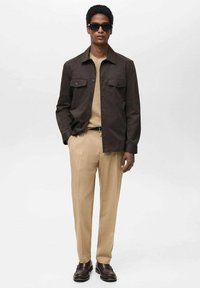 Brun lättviktsjacka med två bröstfickor, beige strukturerad topp, beige byxor och mörka skor. Modell bär mörka solglasögon.