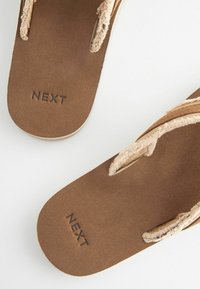 Brūnas flip-flops ar mīkstu, zamša līdzīgu tekstūru, ar beigu auduma siksnām un reljefotu "NEXT" logotipu uz zolītes.