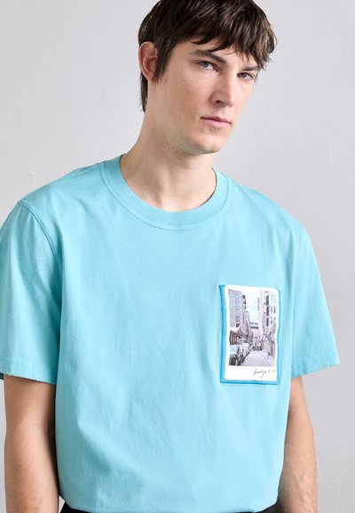 Svijetloplava pamučna majica s džepom na prsima s fotografskim otiskom ulične scene, kontrastni šavovi. Kratki rukavi.