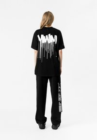 Camiseta negra con un diseño gráfico blanco que presenta gotas y formas angulares en la parte posterior. Combinada con pantalones negros sueltos que muestran texto.