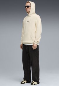Felpa crema con tasca frontale e logo, abbinata a jeans neri larghi e scarpe nere e crema. Modello che indossa occhiali da sole.