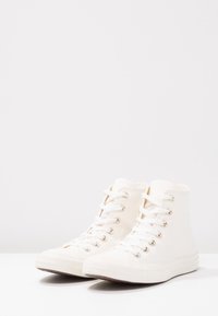 Zapatillas blancas de caña alta hechas de lona, con ojales metálicos, cordones planos, tela texturizada y suela de goma flexible.