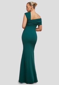 Nainen, jolla on vaaleat hiukset nutturalla, pukeutuneena tehosteväriin fitted off-shoulder-mekkoon, poseeraa kyljellään niin, että selkä näkyy yksinkertaista taustaa vasten.