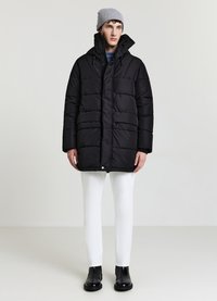 Giubbotto imbottito nero con texture trapuntata, colletto alto e tasche frontali, indossato sopra un maglione blu e pantaloni bianchi. Berretto grigio e stivali neri.